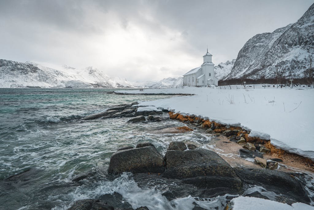 Serene winter scene of a church by the coast in Gimsøysand, Norway, with snow-covered mountains.